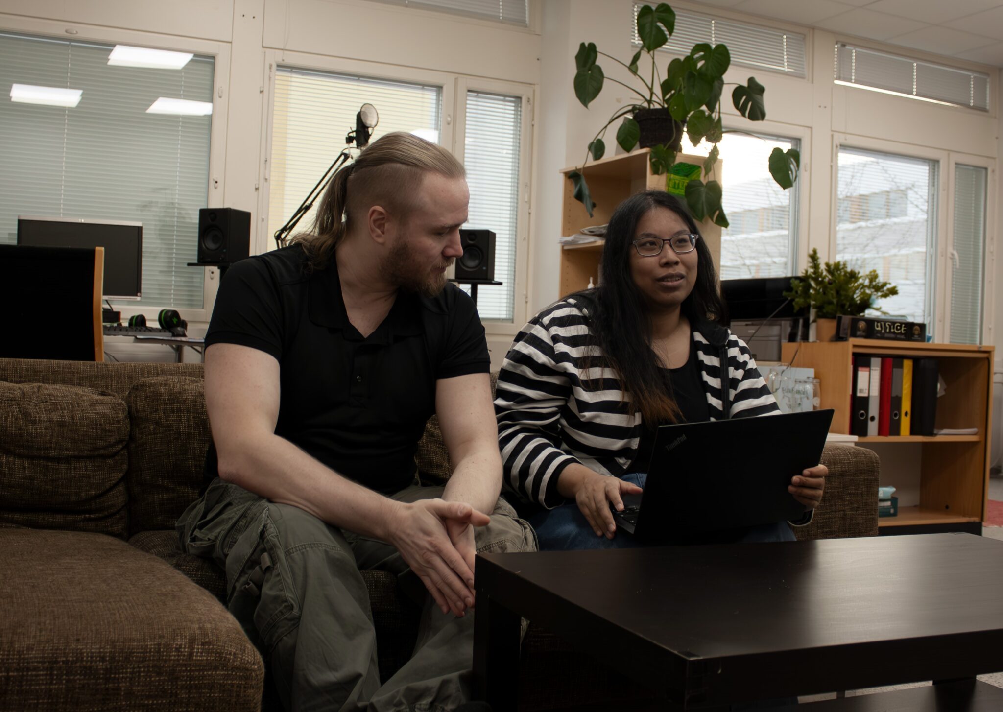 Kuvassa poninhäntäinen mies ja pitkähiuksinen nainen istuvat vierekkäin sohvalla. Mies katsoo nuorta naista, joka katsoo eteenpäin. Edessä olevalla pöydällä naisella on auki oleva kannettava tietokone. Ympärisö on toimistomainen. Sohvan vieressä on iso metallinen patteri ja ikkunan edessä verhot.