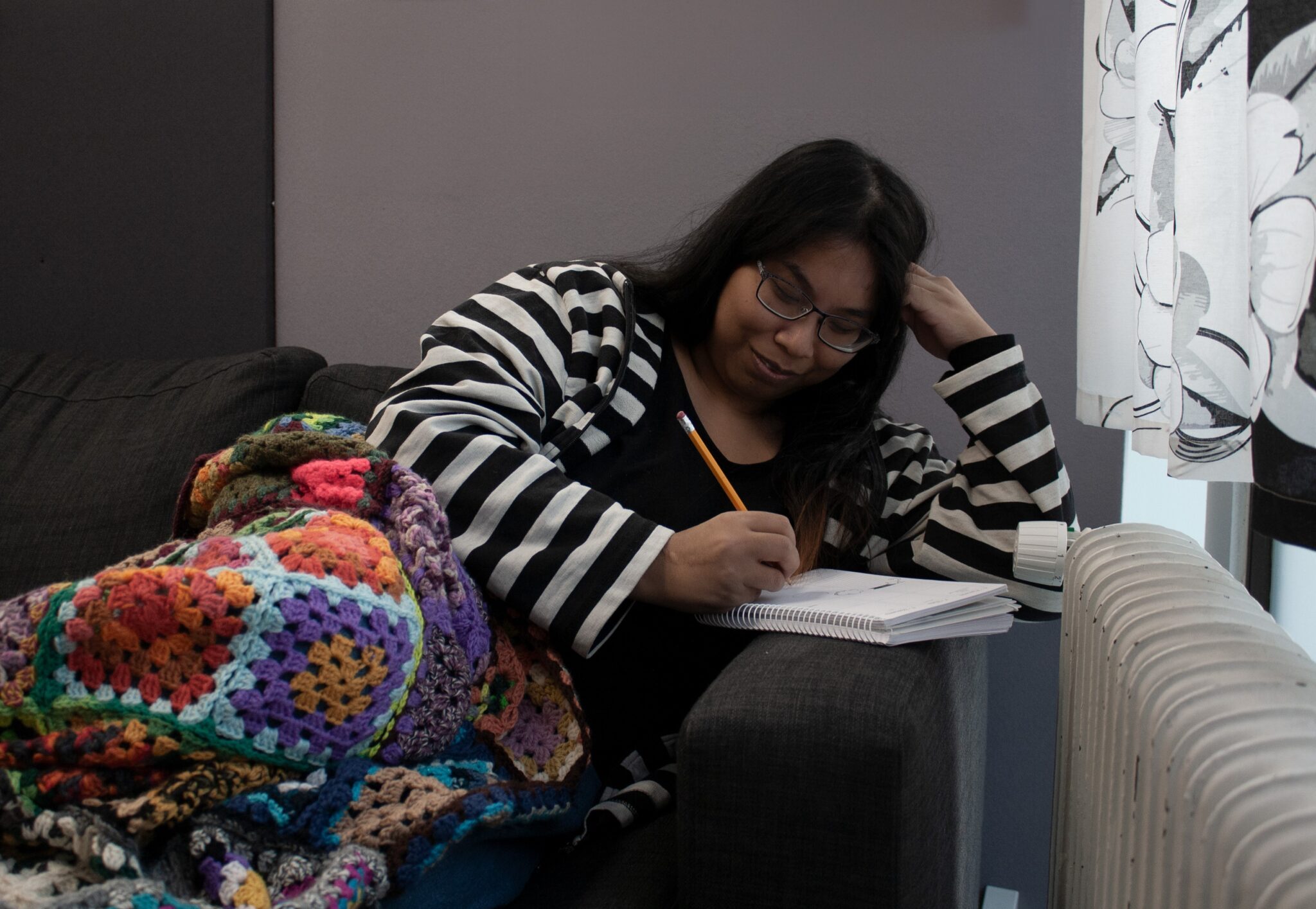 Kuvassa silmälasipäinen nainen pitkissä hiuksissa lojuu rennosti sohvalla neuleshaali jalkojensa päällä. Nainen nojaa kädensijaan ja käsi nojaa päähän, toisella kädessä lyijykynä ja nainen piirtää kädensijan päällä olevaan lehtiöön.