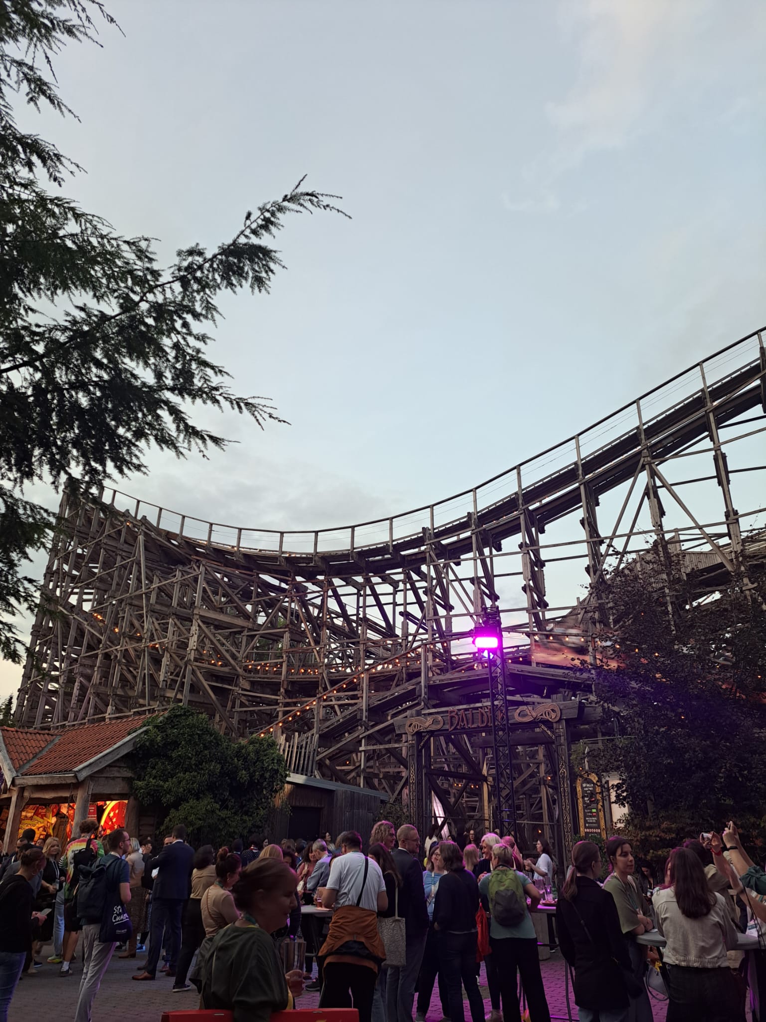 A crowd of people gathered in front of a large wooden roller coaster at an amusement park during the evening, with lights glowing on the structure and trees framing the scene.