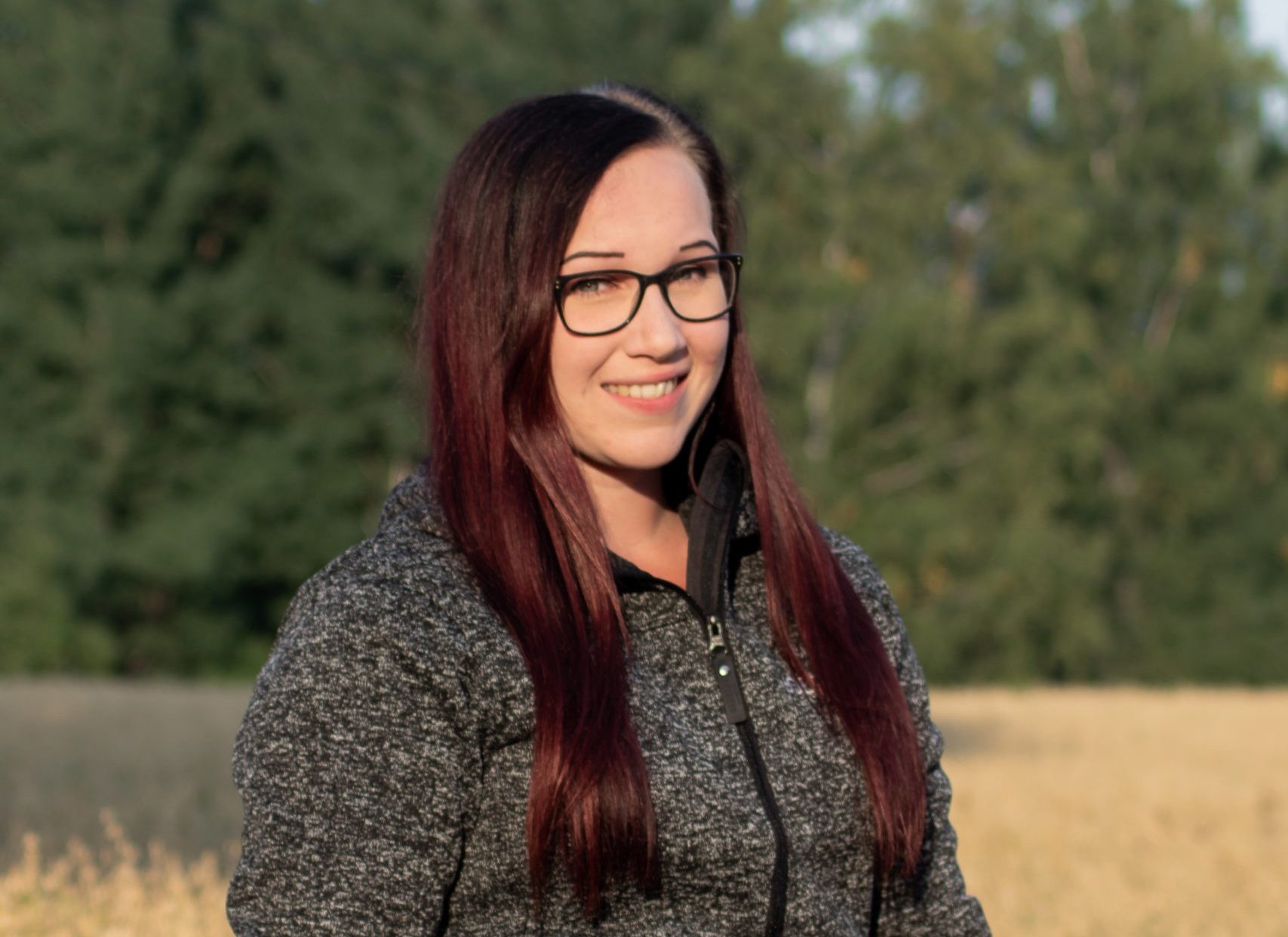 Kuvassa Saija hymyilee. Saijalla on pitkä tumma tukka, harmaa paita ja hän seisoo heinäpellolla.