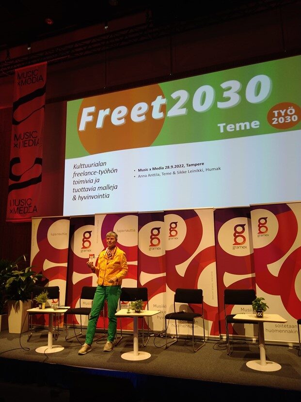 Nainen lavalla, takana powerpoint esitys, jossa kulttuurialan freelance-työhön toimivia ja tuottavia malleja & hyvinvointia otsikko sekä tekstit Freet2030 ja Teme.