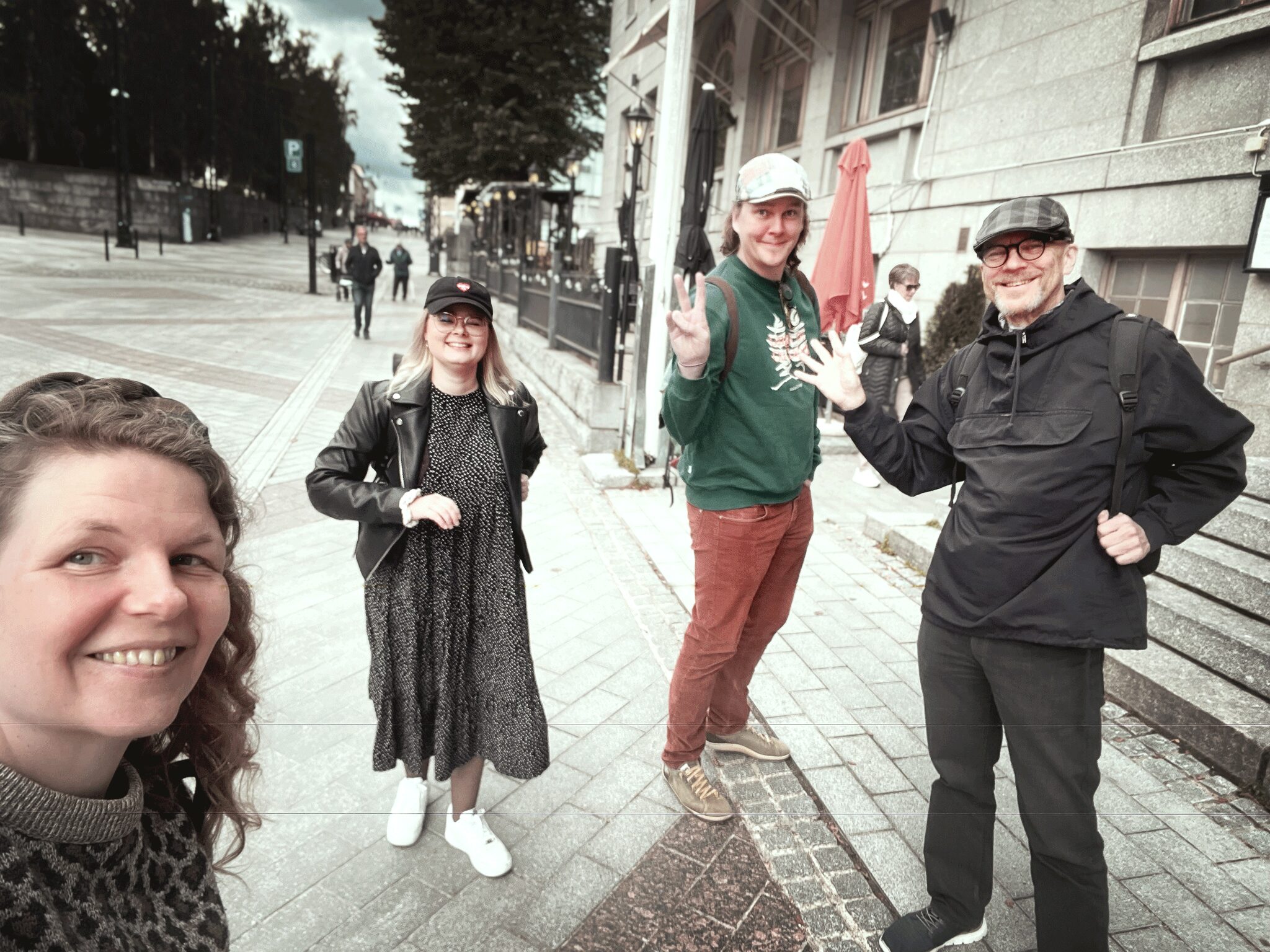 Neljä ihmistä hymyilee ja poseeraa kadulla Kuopiossa tiimipäivän aikana. Taustalla näkyy kivitalo, portaat ja muutamia ohikulkijoita.