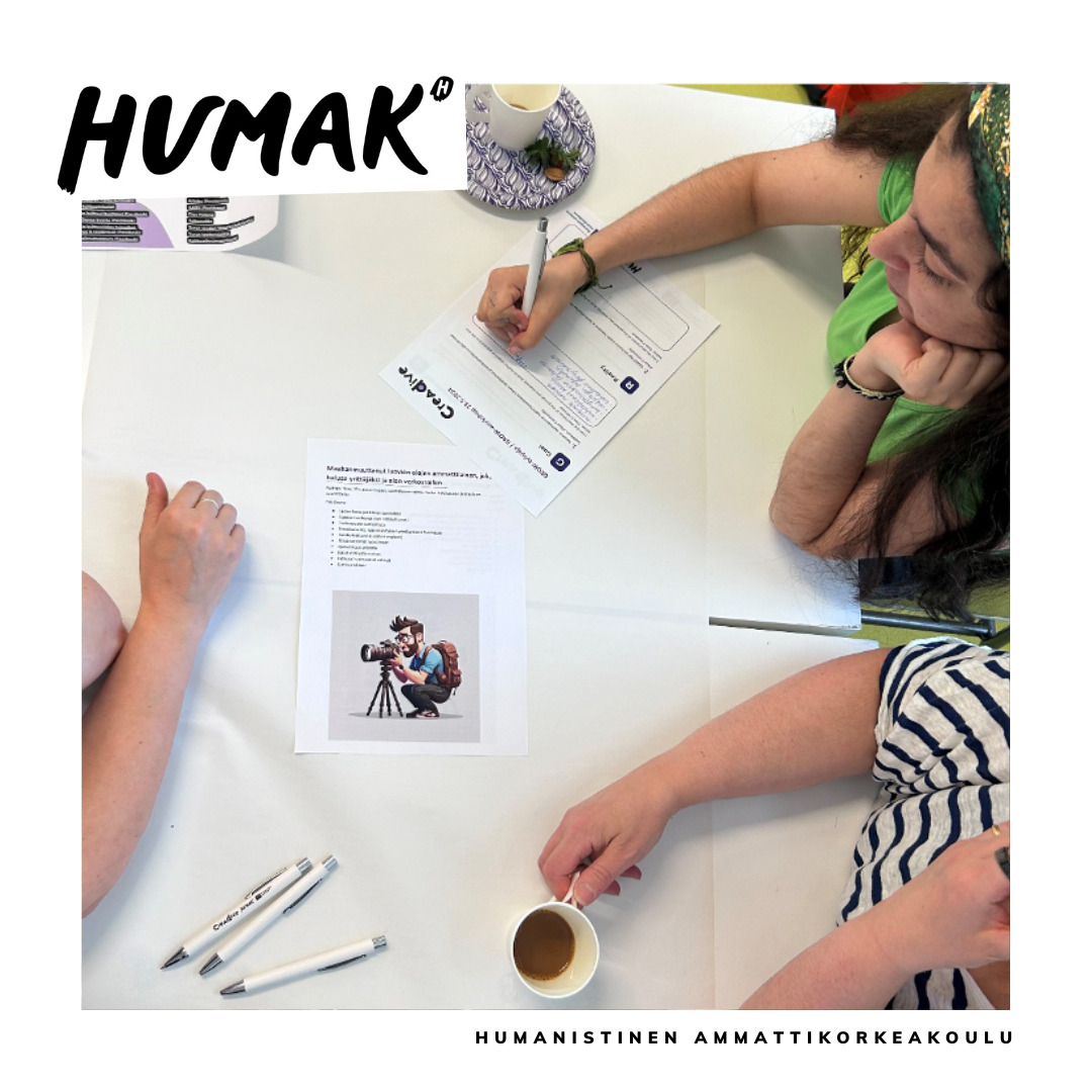 ALT-teksti: Kuvassa näkyy pöytä, jossa on kyniä, papereita ja kolme ihmistä, joista yksi kirjoittaa paperille.