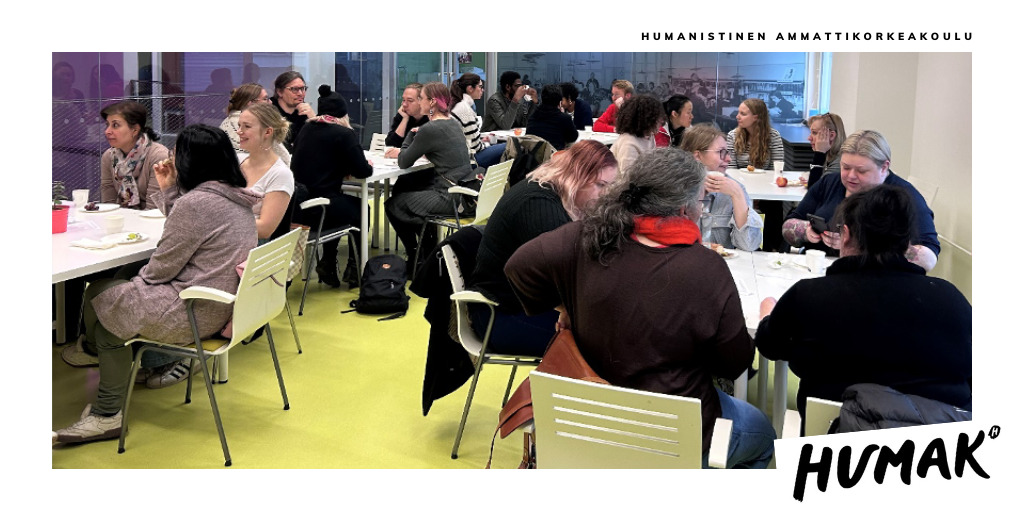 Kuvassa näkyy luokkahuone, jossa viiden pöydän ääressä istuu ihmisiä keskustelemassa.