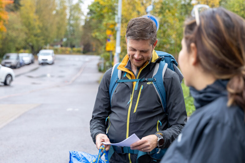 Henkilö seisoo ulkona kallioisella alueella ja pitää vihkoa kädessään, edessä ryhmä kuuntelemassa.