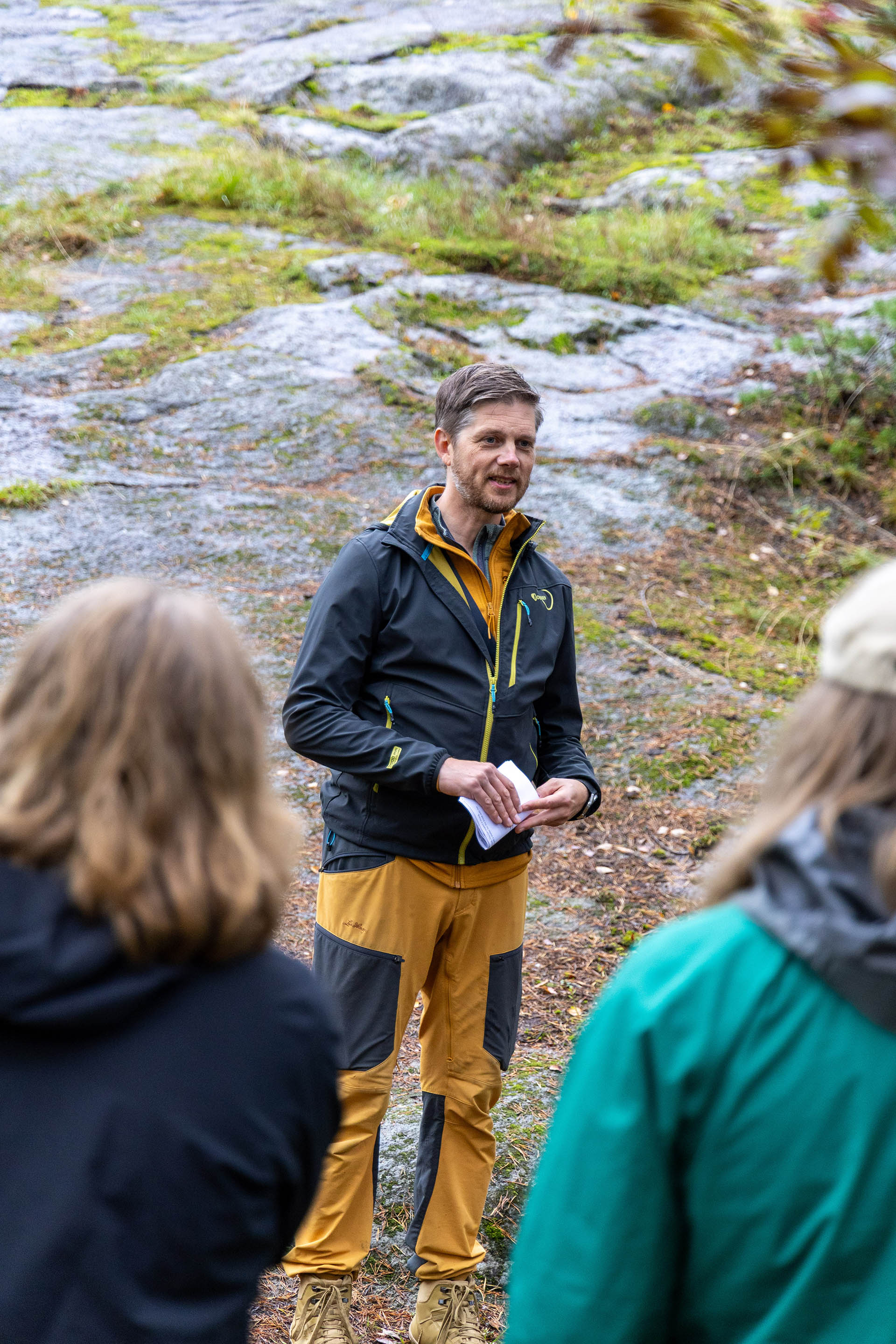 Henkilö seisoo ulkona kallioisella alueella ja pitää vihkoa kädessään, edessä ryhmä kuuntelemassa.