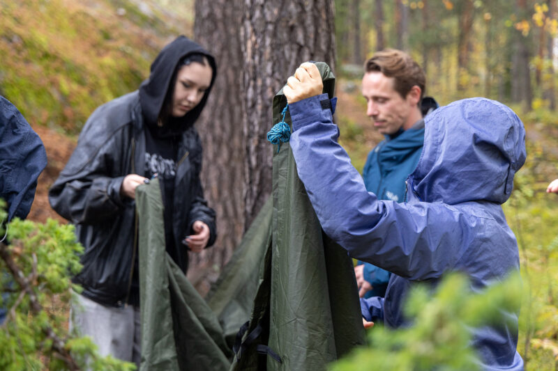 Kolme henkilöä pitää telttakangasta metsässä, taustalla näkyy puu ja syksyisiä lehtiä.