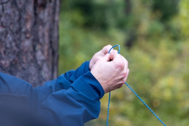 Lähikuva käsistä, jotka sitovat sinistä narua puun ympärille metsäisessä ympäristössä.