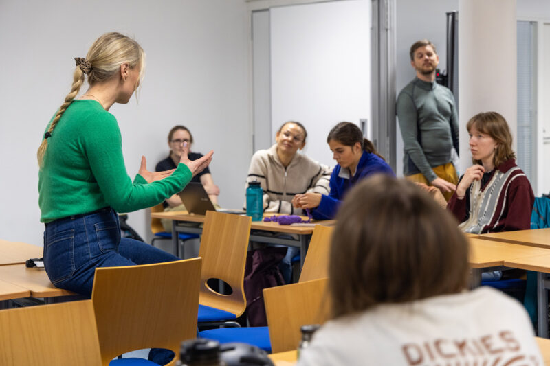 Henkilö seisoo luokkahuoneessa ja keskustelee muiden kanssa, jotka istuvat pöytien ääressä.