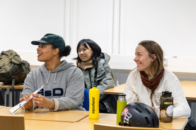 Kolme henkilöä istuu luokkahuoneessa pöytien äärellä ja nauraa.
