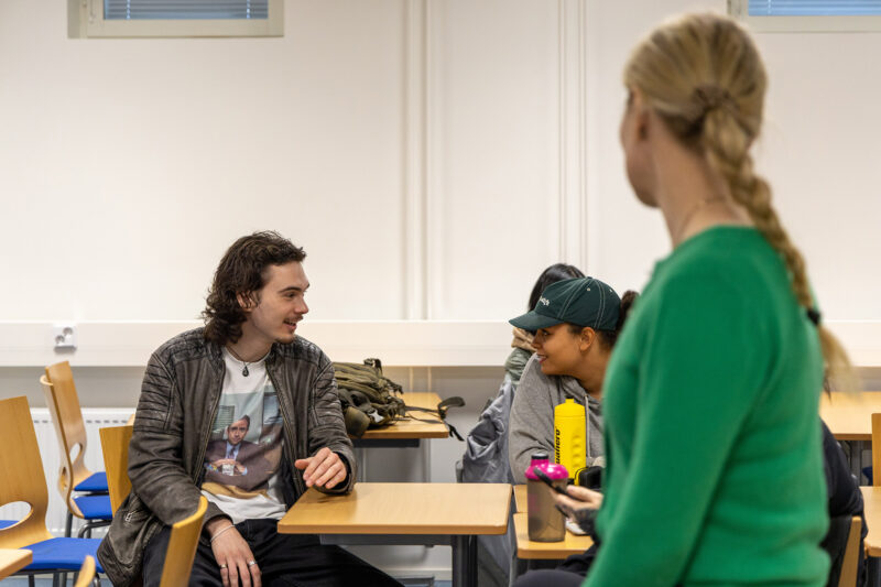 Kaksi henkilöä istuu luokkahuoneessa pöydän ääressä, edessä vesipulloja ja varusteita. Etualalla sumea henkilö.