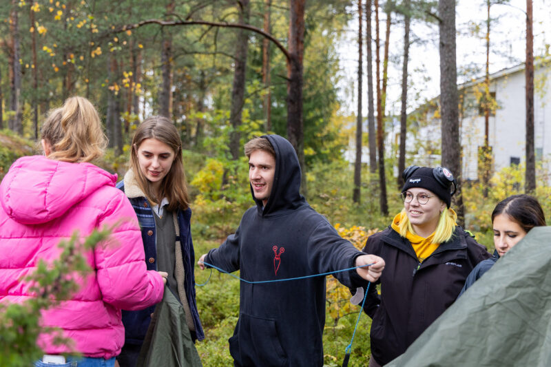 Ryhmä ihmisiä seisoo metsässä ja pitää telttakangasta ja naruja, valmistellen majoitteen pystytystä.