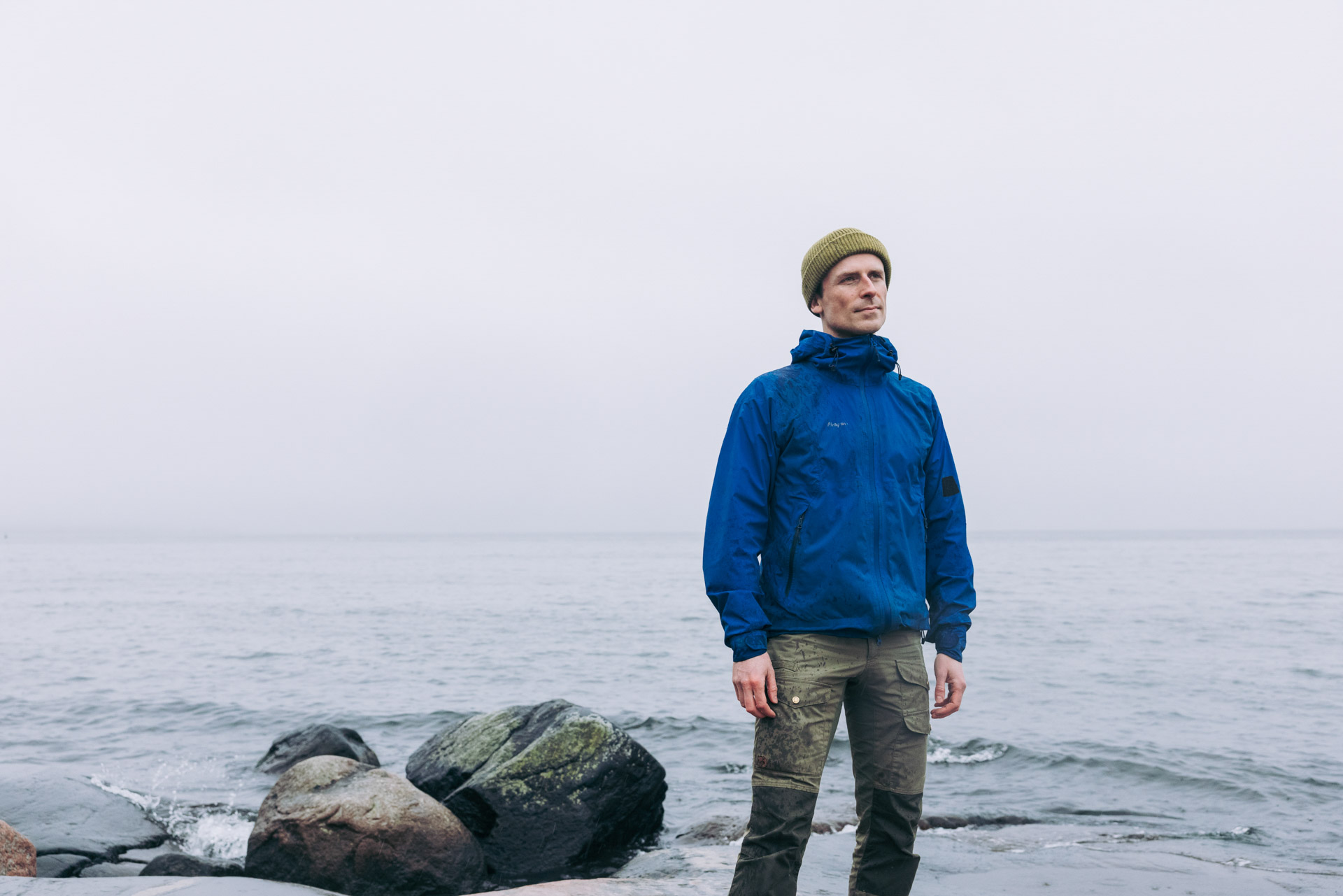 Henkilö seisoo meren rannalla kivikkoisessa maisemassa sumuisessa säässä.