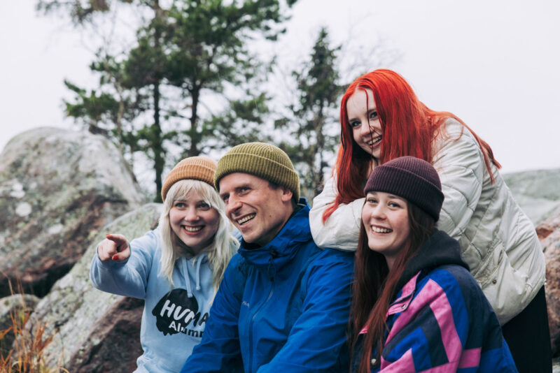 Neljä henkilöä istuu kallioilla metsäisessä ympäristössä ja katsoo samaan suuntaan. Vasemmanpuolinen henkilö osoittaa eteenpäin.