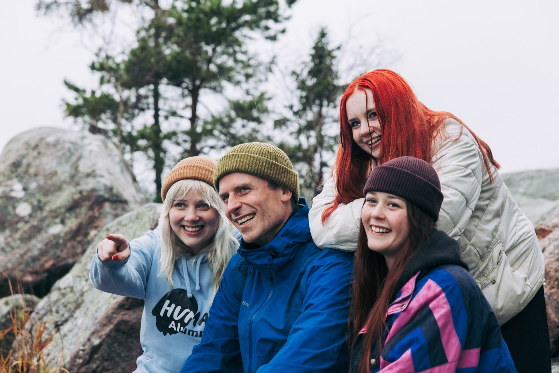 Neljä henkilöä istuu kallioilla metsäisessä ympäristössä ja katsoo samaan suuntaan. Vasemmanpuolinen henkilö osoittaa eteenpäin.