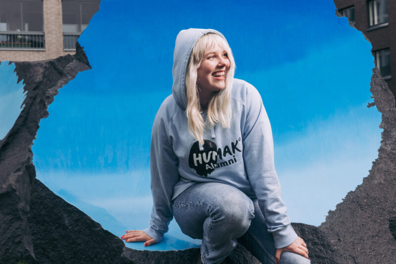 Yksittäinen henkilö istuu siniseksi maalatun kiven reunalla. Taustalla näkyy kaupunkiympäristöä. Henkilö hymyilee ja hänellä on päällään "Humak Alumni"-huppari.