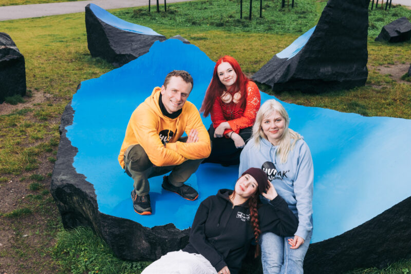 Neljä henkilöä istuu tai nojaa siniseksi maalattuun kallioon puistomaisessa ympäristössä,