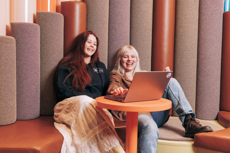 Kaksi henkilöä istuu sohvalla ja työskentelevät yhdessä kannettavan tietokoneen äärellä, joka on pöydällä. Henkilöt nauravat.