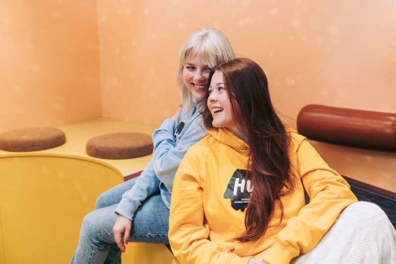 Kaksi henkilöä istuu rennosti sisätilassa värikkäiden kalusteiden keskellä ja nojaavat toisiinsa. Henkilöillä on päällään Humak-hupparit.