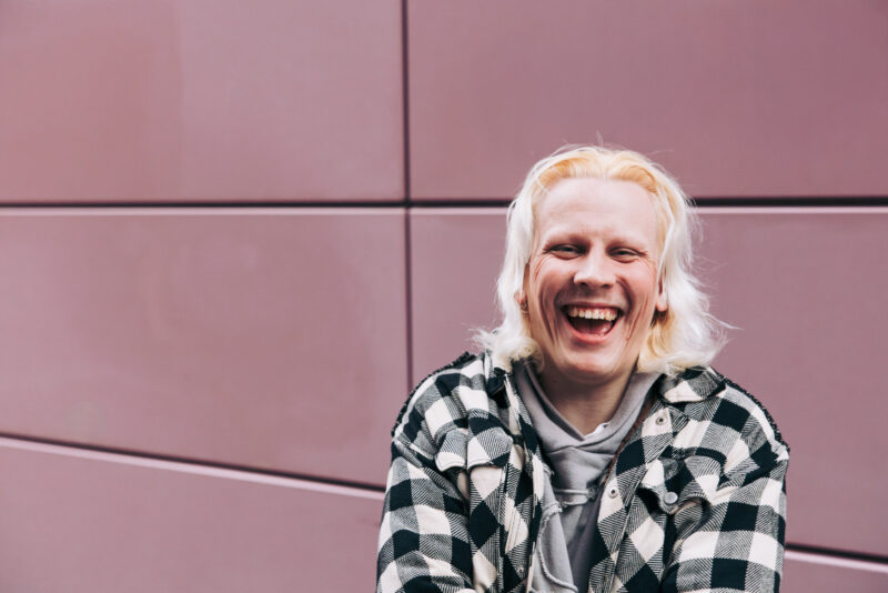 Puolikuva henkilöstä, joka seisoo vaaleanpunaisen seinän edessä ja katsoo eteenpäin nauraen.