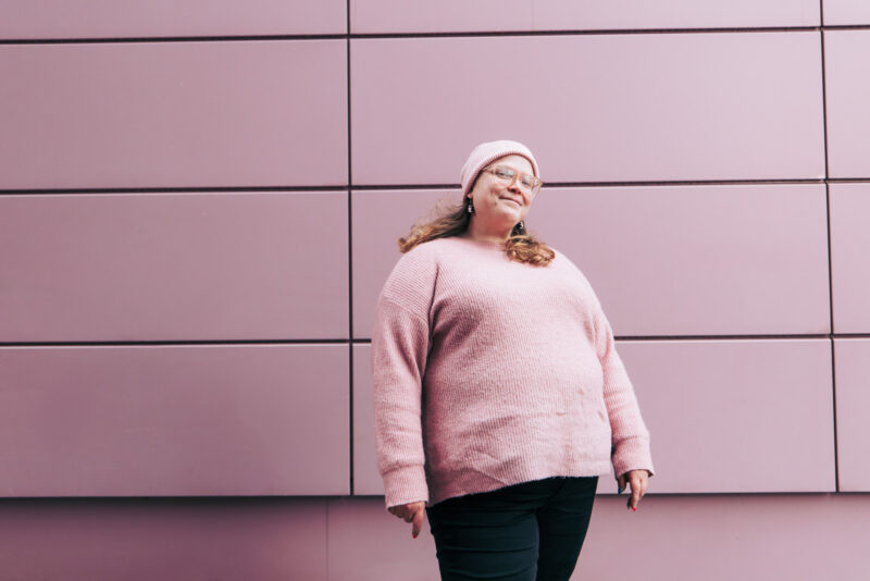 Henkilö seisoo vaaleanpunaisen, geometrisen seinän edessä ulkotilassa. Henkilöllä on päällään vaalean punainen pipo, vaaleanpunaiset lasit, vaaleanpunainen paita ja mustat housut.