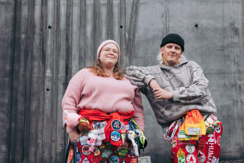 Kaksi henkilöä seisoo betoniseinää vasten ja hymyilevät hieman. Molemmilla on yllään punaiset opiskelijahaalarit, jotka ovat täynnä erilaisia haalarimerkkejä.