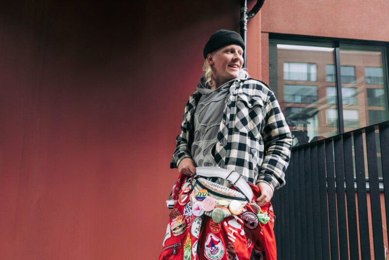 Ulkona rakennuksen edessä seisova henkilö, jolla on yllään punaiset opiskelijahaalarit, jotka ovat koristeltu haalarimerkeillä. Henkilö hymyilee ja katsoo oikealle.