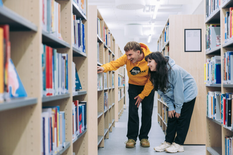 Kaksi henkilöä selaa kirjoja kirjaston hyllyjen välissä, kumartuneina tutkimaan alemman hyllyn valikoimaa. Henkilöillä on päällä Humak-hupparit.