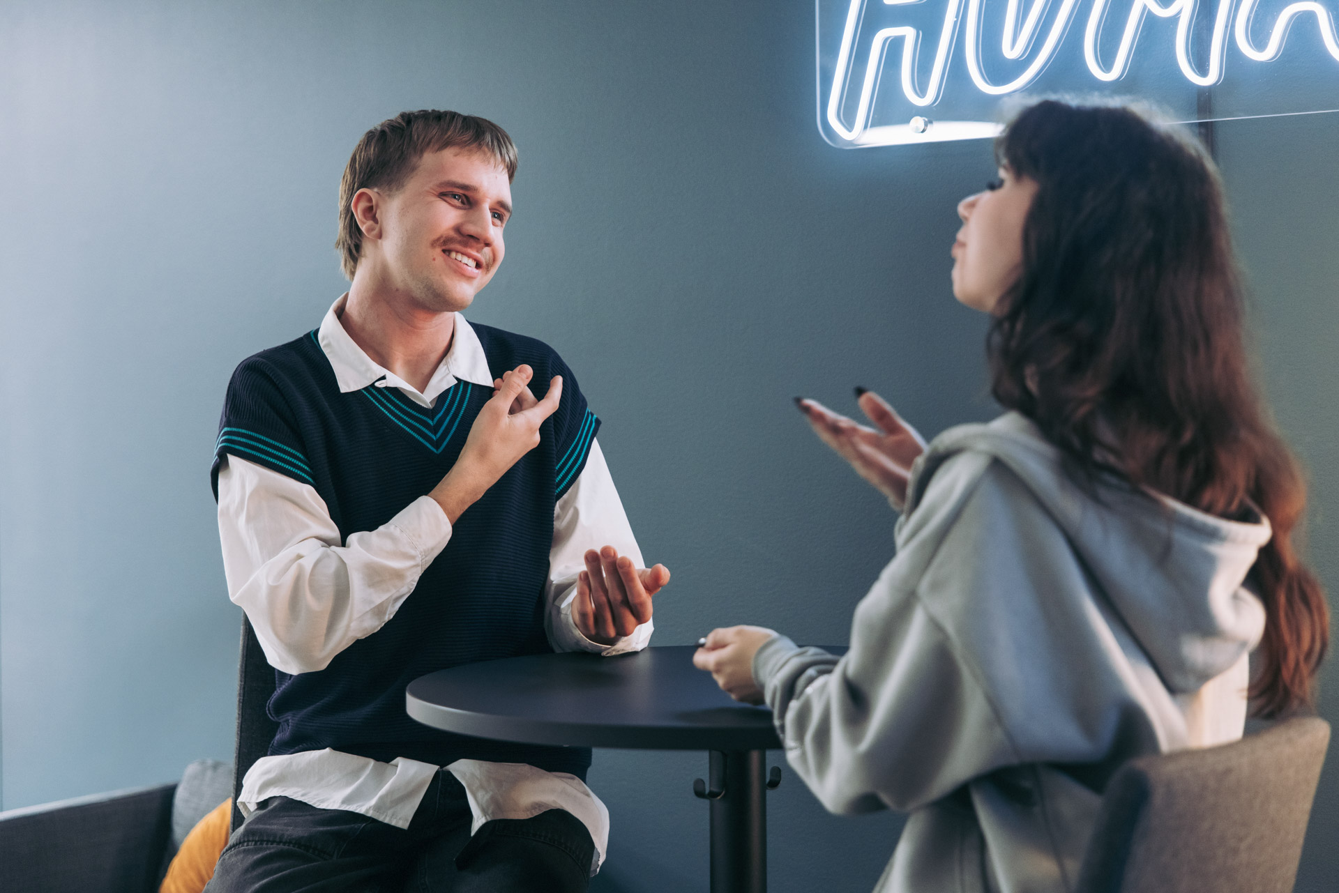 Kaksi henkilöä istuu pöydän ääressä sisätilassa ja keskustelee viittomakieltä käyttäen.