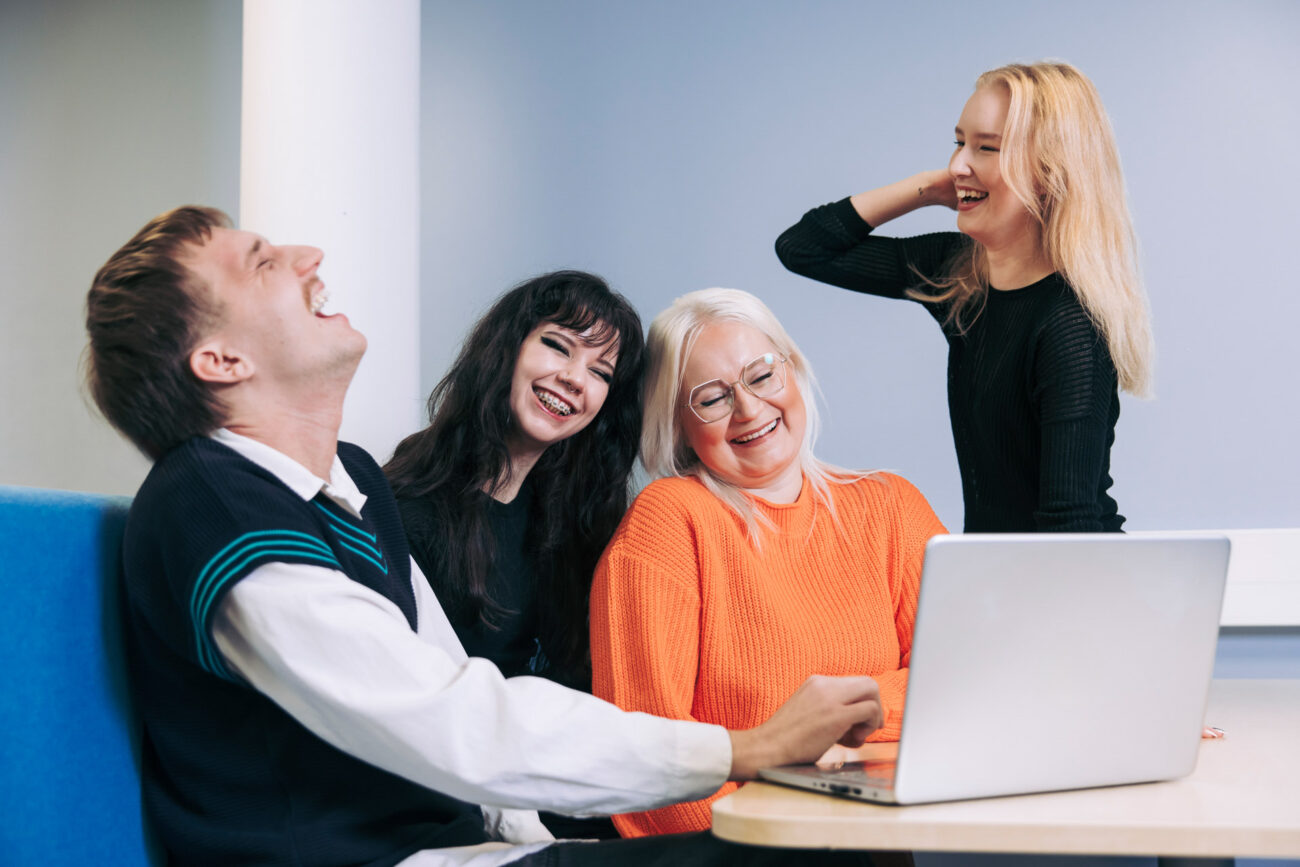 Neljä henkilöä istuu pöydän ympärillä ja nauravat. Henkilöt katsovat yhdessä kannettavan tietokoneen näyttöä sisätilassa.