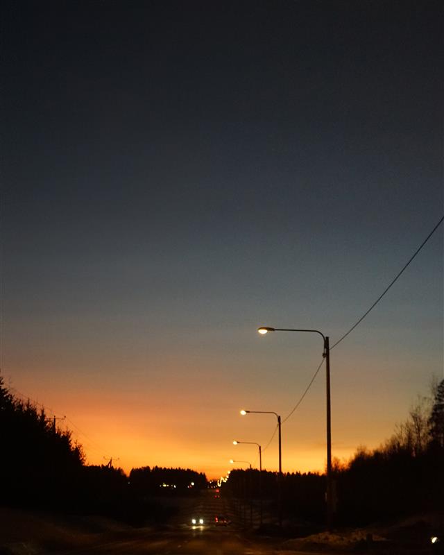 Autotie ja katuvalot auringonlaskun aikaan. Tumma metsänreuna ja pimenevä taivas.
