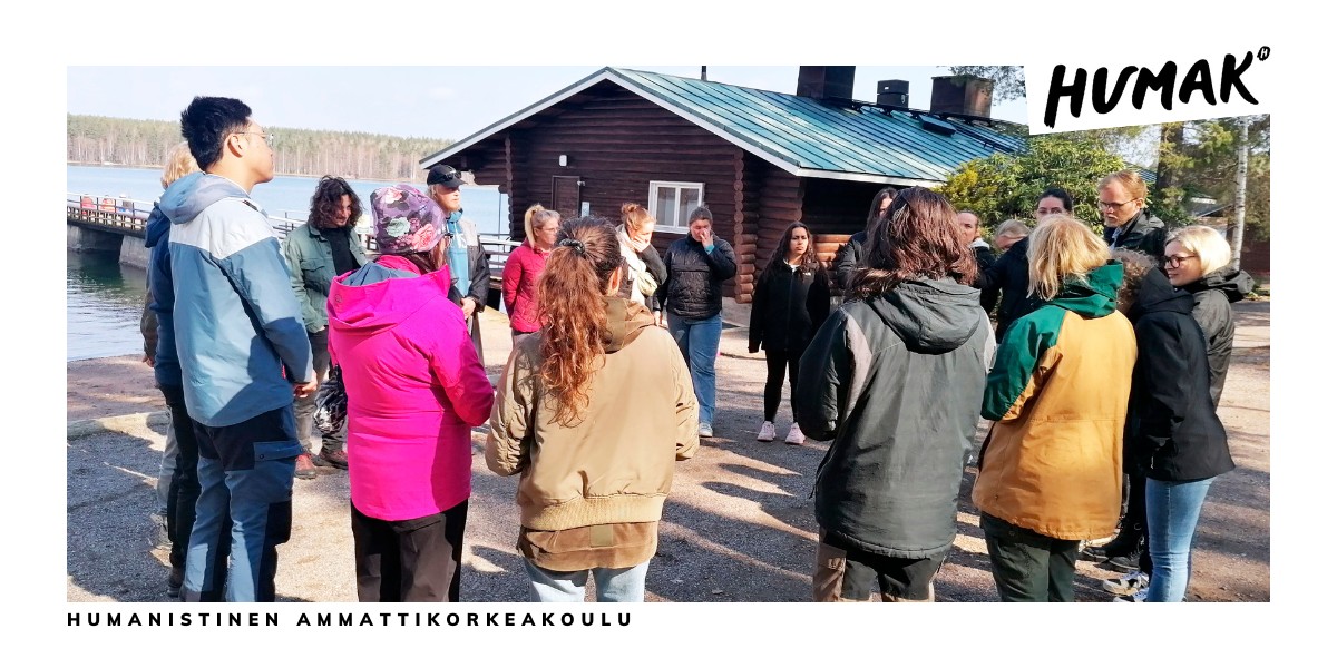 Opiskelijoita eri kansallisuuksista seisoo ringissä järven rannalla.