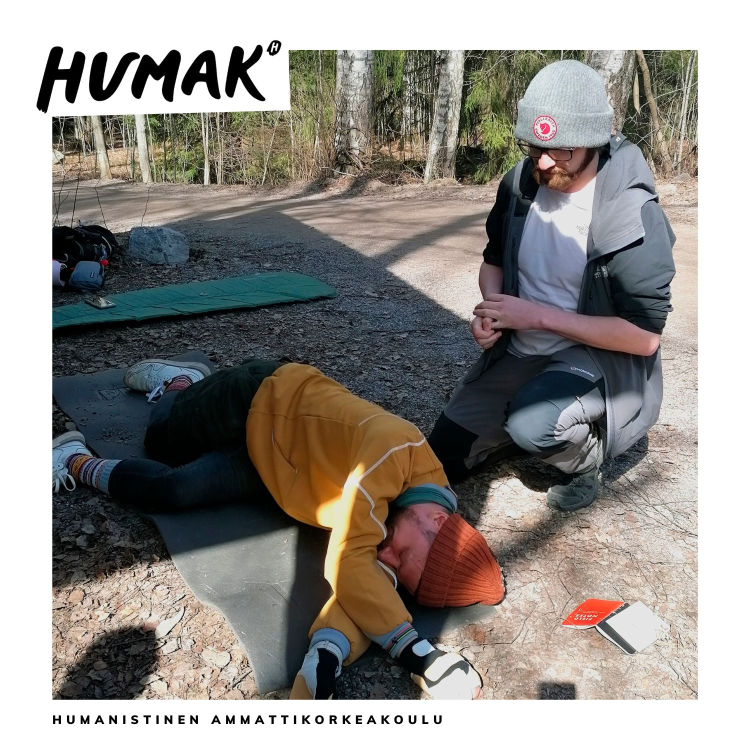 Luontoympäristössä toinen opiskelija makaa maassa kylkiasennossa ja toinen opiskelija on polvistunut hänen taaksensa tarkastamaan hänen vointiaan.