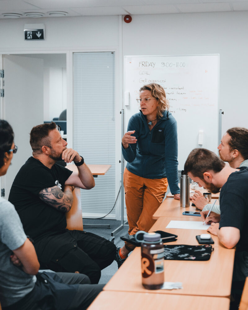 Henkilö seisoo luokkahuoneessa pöytien ääressä ja selittää jotakin muille, jotka istuvat ja kuuntelevat. Taustalla on valkotaulu, jossa näkyy tekstiä ja aikataulu.