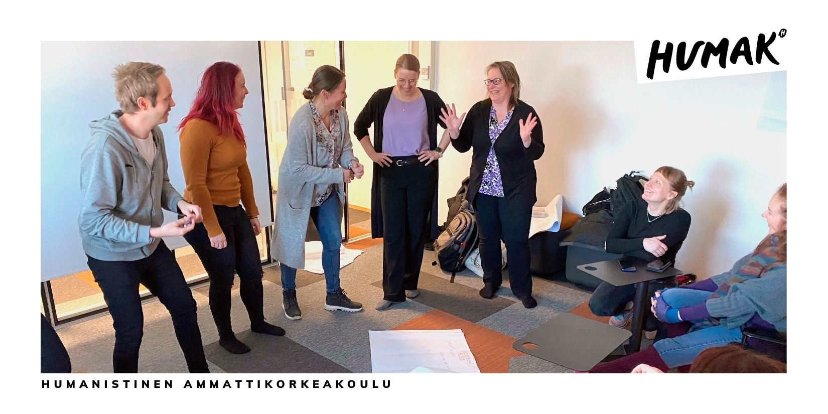 Viisi henkilöä seisoo rivissä luokkahuoneessa. Neljäs vasemmalta seisoo huvittuneena kädet puuskassa ja muut katsovat häntä hymyillen. 