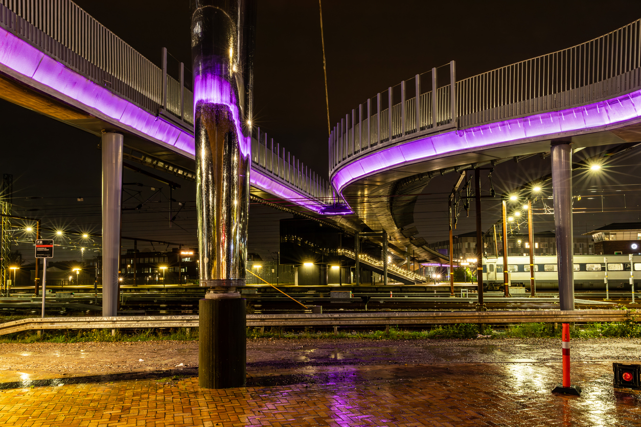 Valokuva öisestä ja sateisesta kaupungista. Kuvan yläosassa on kaksi kaareilevaa raidetta, joita koristaa violetti valoelementti. 