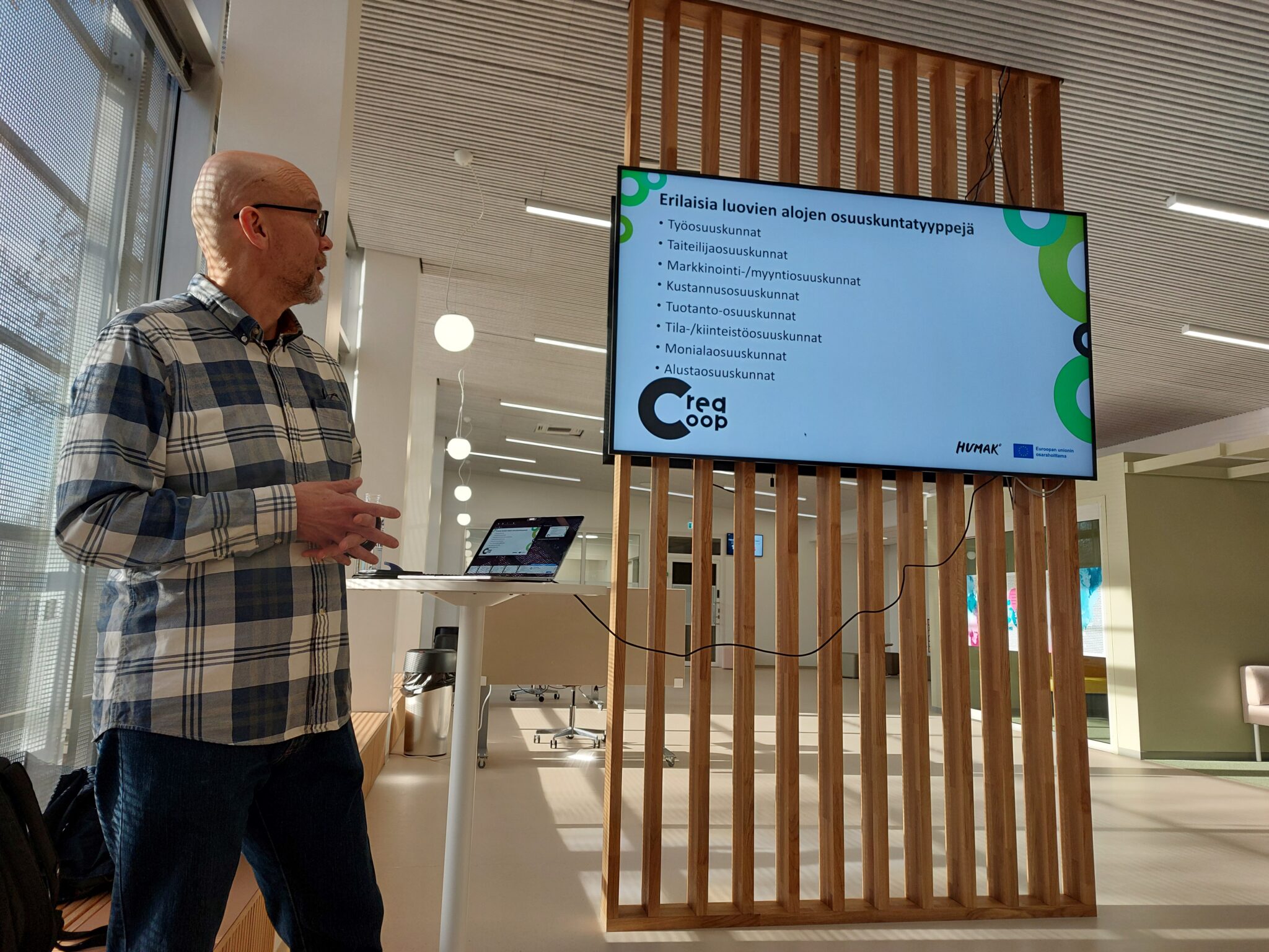 Henkilö seisoo sisätilassa suuren näytön vieressä, jossa näkyy dia otsikolla 'Erilaisia luovien alojen osuuskuntatyyppejä'. Näytöllä lueteltu useita osuuskuntatyyppejä, kuten työosuuskunnat, taiteilijaosuuskunnat ja markkinointi-/myyntiosuuskunnat. Vieressä korkea pöytä ja kannettava tietokone.