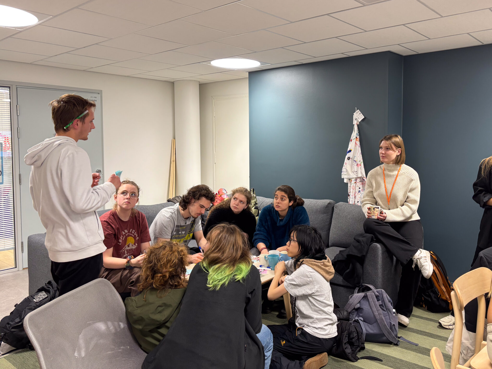 A group of students discussing on couches. Others listen to one student speaking.
