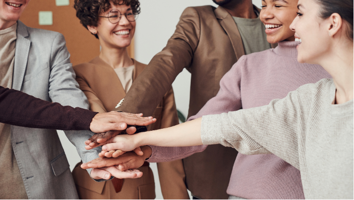 Viisi ihmistä ojentaa oikeat kätensä keskelle, muodostaen kämmenistä päällekkäin asetetun pinon. Ihmisten ihonvärit ovat erilaisia, mikä tuo esiin moninaisuuden ja yhteisöllisyyden.