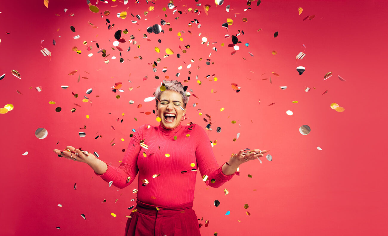 Punaisen taustan edessä punaisiin pukeutunut lyhythiuksinen nainen pitää käsiään ylhäällä ja nauraa leveästi. Hänen päällensä lentää erivärisiä konfetteja.