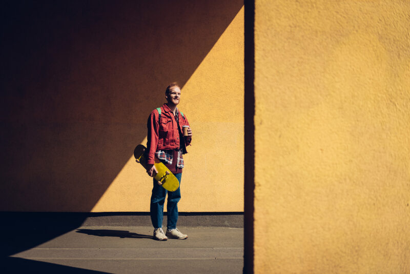 Henkilö seisoo kellertävän betoniseinän edessä. Hän pitää kädessään skeittilautaa sekä kahvikuppia.