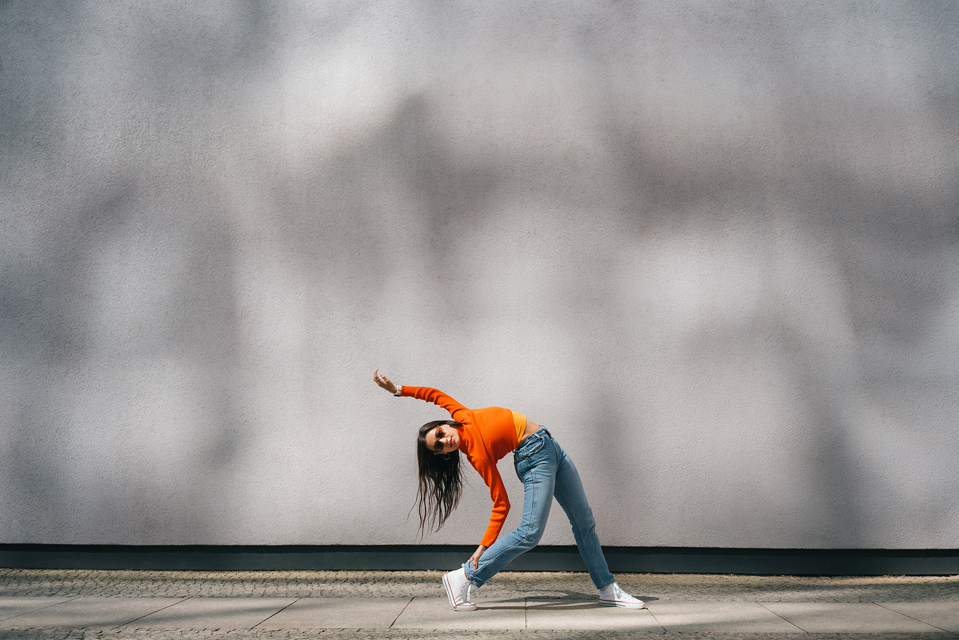 Harmaa betonitausta, jonka edessä oranssipaitainen nainen venyttää itseään.