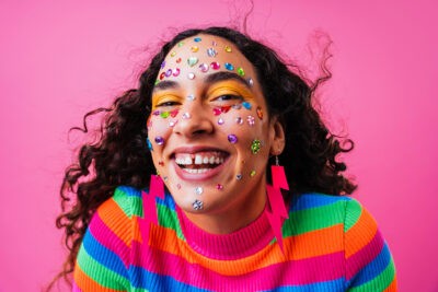 Nainen hymyilee todella leveästi kameralle. Hänellä on päällään monivärinen paita, salamanmuotoiset pinkit korvakorut sekä hänen naamaansa on liimattu timanttitarroja.