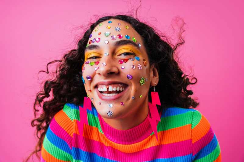 Nainen hymyilee todella leveästi kameralle. Hänellä on päällään monivärinen paita, salamanmuotoiset pinkit korvakorut sekä hänen naamaansa on liimattu timanttitarroja.