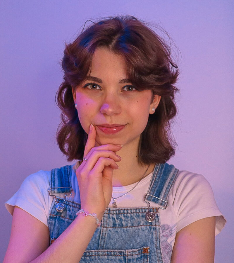 Henkilö seisoo violettisävyisessä valaistuksessa, pukeutuneena farkkuhaalareihin ja vaaleaan t-paitaan. Hän pitää kättään kevyesti leuallaan pohtivassa asennossa.