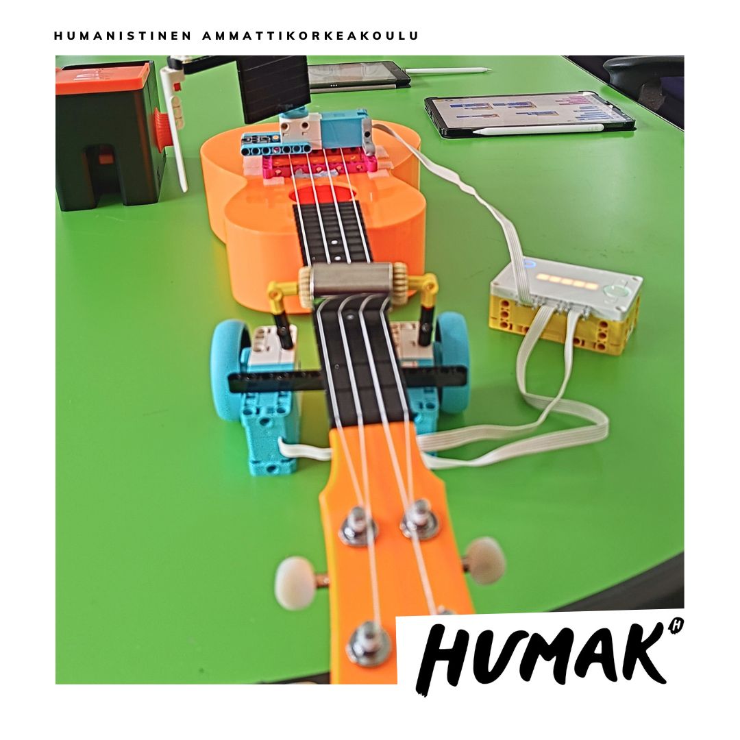 Lähikuva oranssista sähköisestä ukulelesta, johon on kiinnitetty pieni valkoinen soittorobotti. 