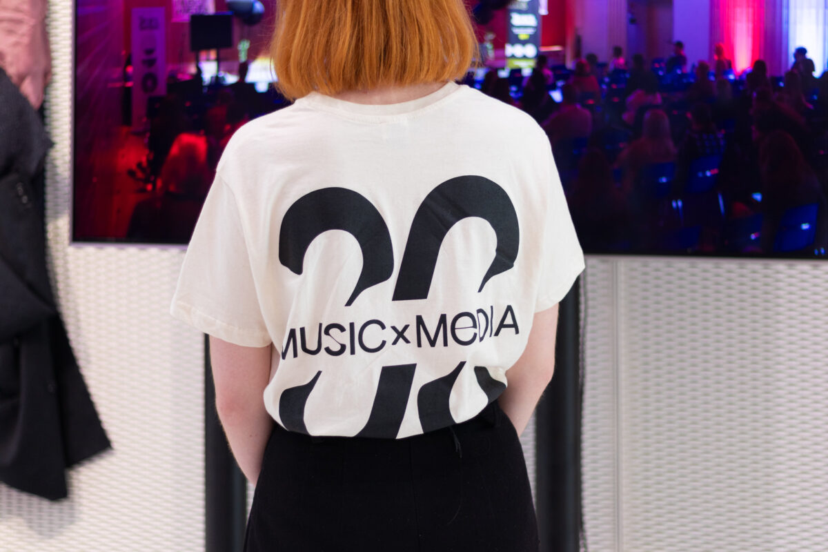 Henkilö poseeraa selkä kohti kameraa, hänellä on päällään valkoinen t-paita, jossa mustalla teksti Music x Media.