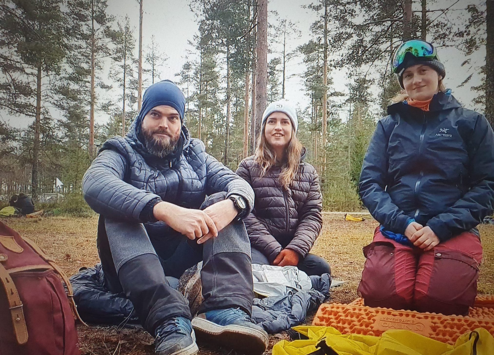 Metsäisessä maastossa, reppujen vieressä istuvat mies vasemmalla, nainen takana keskellä, nainen oikealla. Kaikilla on päällään ulkoiluvaatteet ja päähineet. Oikeanpuolisella naisella on päässään pipo ja laskettelulasit.