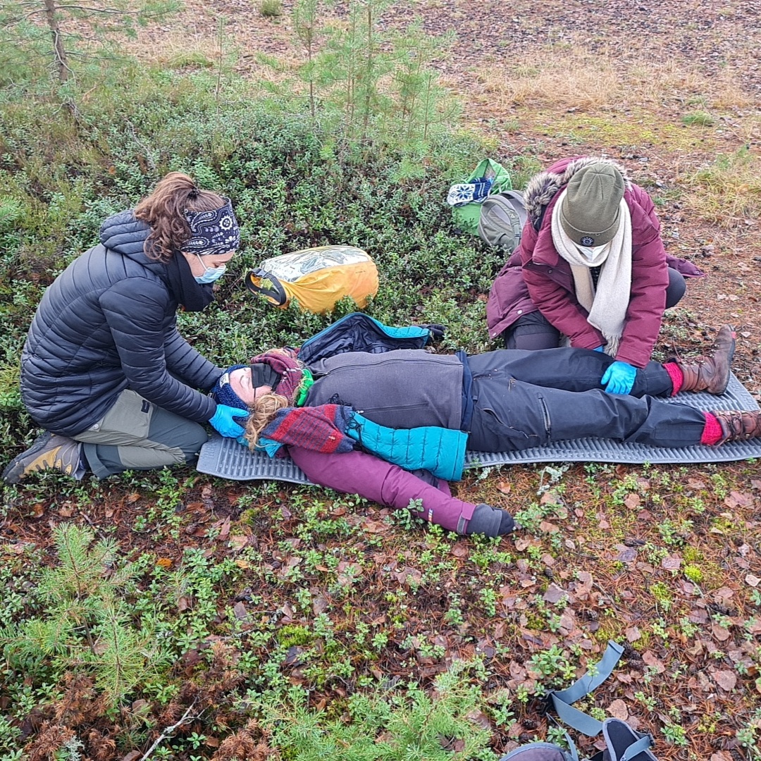 Varpujen päällä makaa nainen ja hänen päätään pitää toinen nainen sekä kolmas nainen pitää kiinni makaavana henkilön jalasta. Kaikki ovat ulkovaatteissa. 