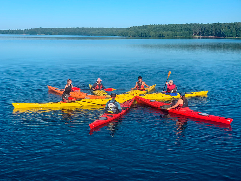 Seven people floating on a lake in kayaks, face-to-face forming a star shape. The lake is calm and there are green forests in the background.